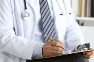 Male medicine doctor hand holding silver pen writing something on clipboard closeup. Ward round, patient visit check, medical calculation and statistics concept. Physician ready to examine patient